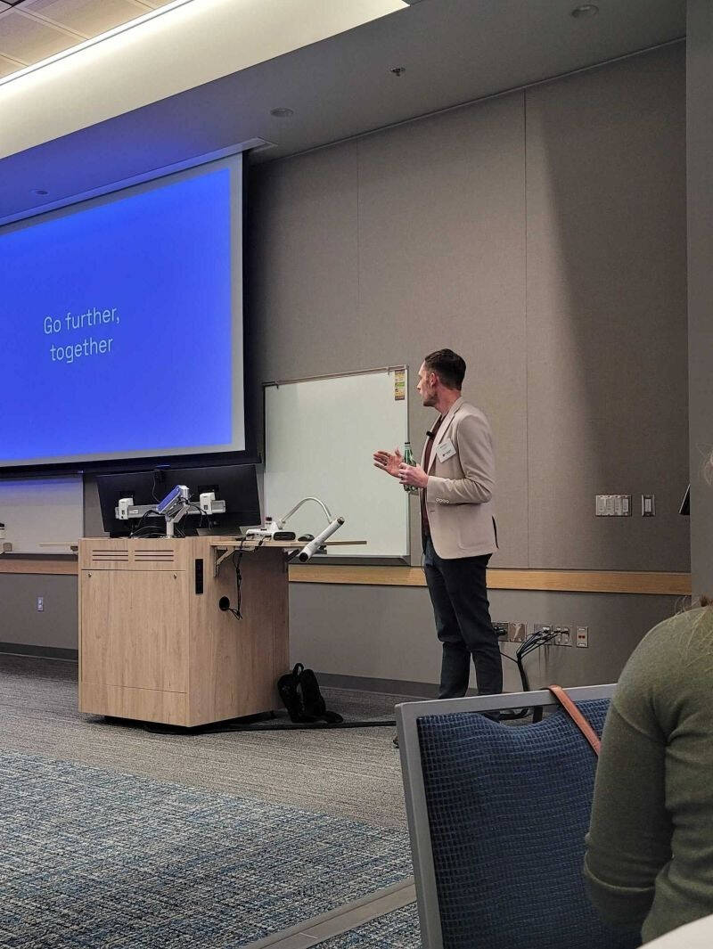 A person in business attire presents at the front of a modern classroom, gesturing while speaking. The projection screen behind them reads “Go further, together,” and nearby are a lectern, computer equipment, and a backpack on the floor.